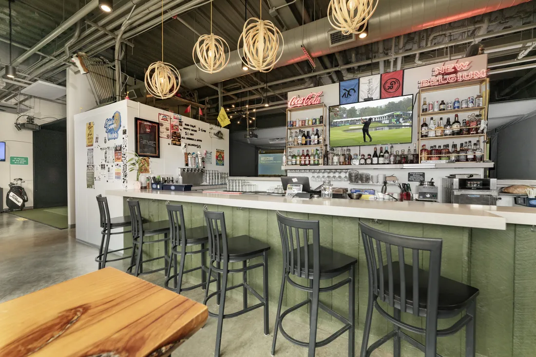 The Birdie Lounge bar: stools along a green counter, bottles and a golf broadcast behind.