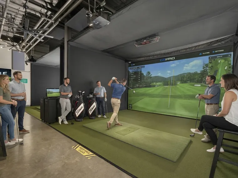 Friends watching one of their group swing in a Birdie Lounge bay, the fairway up on the big screen.